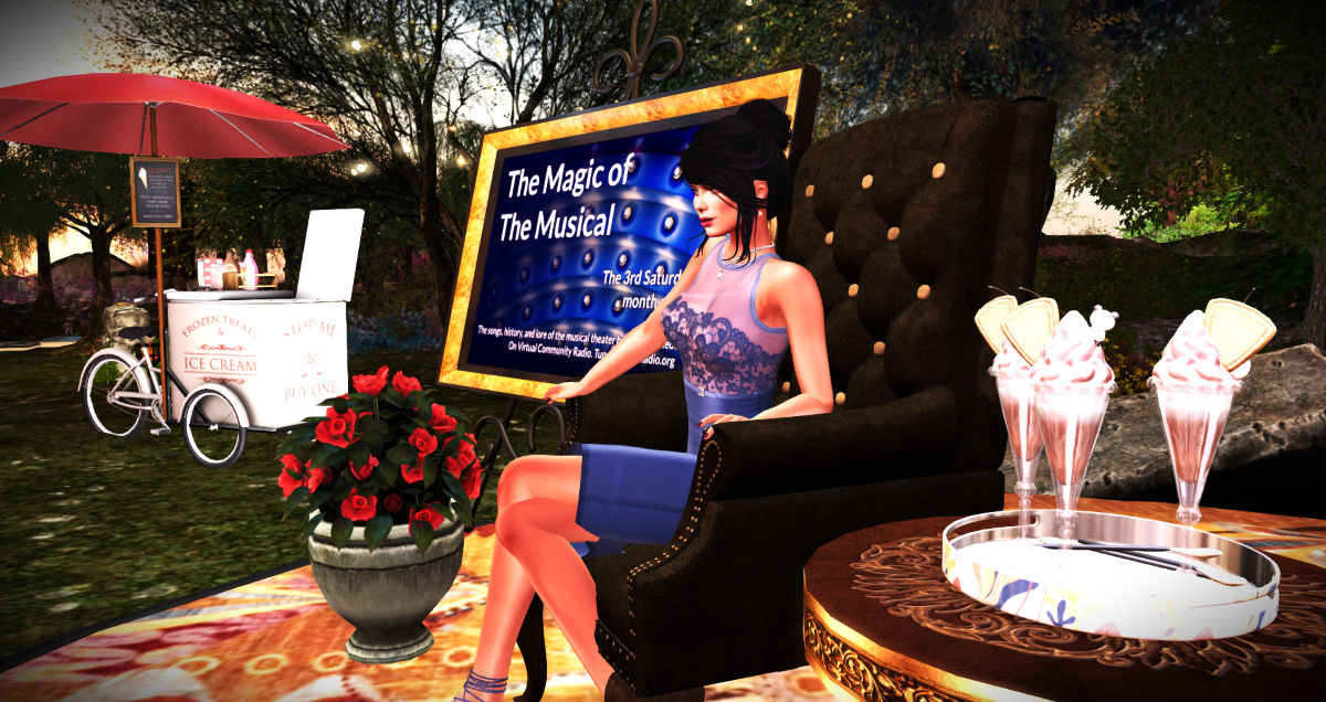 Host (female - kight skinned) in a blue dress with dark brown hair, seated in front of a poster for The Magic if The Musical